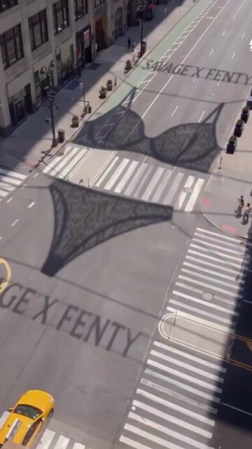 A Savage X Fenty aproveitou a New York Fashion Week para criar uma campanha que simula estruturas gigantes de lingerie suspensas sobre a região de Seventh Avenue e Fashion Avenue, no Garment District de Manhattan, projetando sombras de sutiã e calcinha sobre as ruas.
Apesar da aparência realista, a instalação não existe fisicamente: as imagens são um trabalho de CGI que simula a intervenção da marca nas ruas de Nova York.