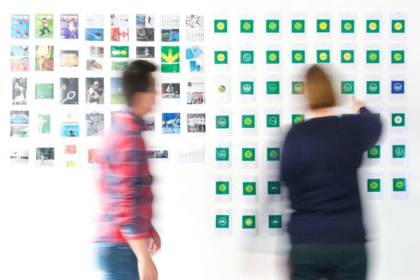 Duas pessoas organizando post-its em um mural branco durante sessão de brainstorming, notas em verde e amarelo alinhadas em colunas.