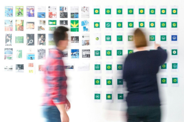 Duas pessoas organizando post-its em um mural branco durante sessão de brainstorming, notas em verde e amarelo alinhadas em colunas.