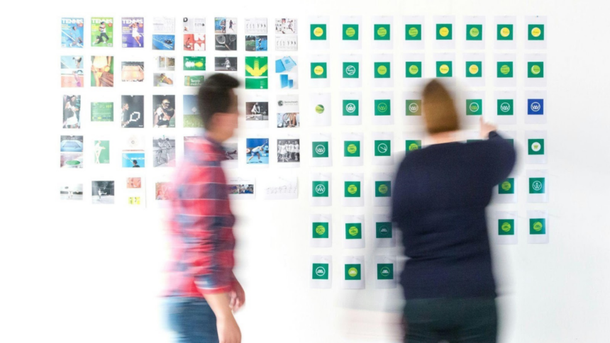 Duas pessoas organizando post-its em um mural branco durante sessão de brainstorming, notas em verde e amarelo alinhadas em colunas.