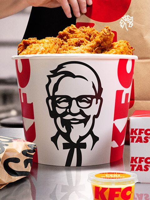 O fast-food sempre disputou dois públicos: a família que quer praticidade e o jovem que quer experiência, onde o KFC tomou uma decisão e escolheu um lado da história. A reformulação reorganiza produto, identidade e espaço físico em torno de um consumo individual, personalizado e digital.
O logo ganhou letras mais cheias e cantos arredondados para funcionar num ícone de aplicativo. As embalagens chegam com frases em vermelho e preto feitas para ser fotografadas. O cardápio estreia a Kwench, linha de bebidas com milkshake, limonada e café gelado, ao lado de mais de 20 molhos adaptáveis por mercado.
A estratégia é clara: sair do balde compartilhado no centro da mesa e entrar no pedido individual que o cliente monta, fotografa e posta.